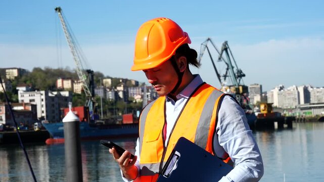 Professional asian port engineer in a safety vest and hard hat managing logistics and talking on his mobile phone while standing in front of large cargo cranes and container ships at a busy harbor