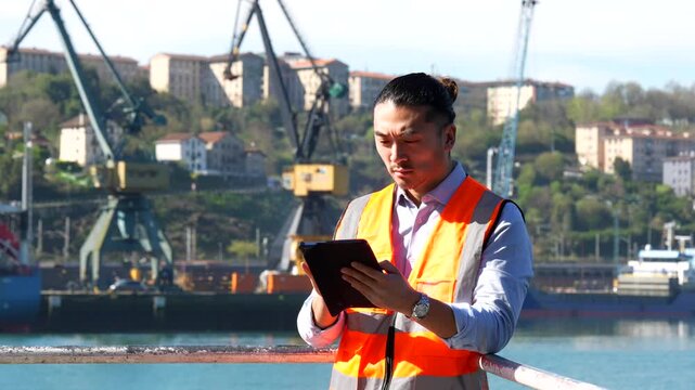 Young asian male dock worker in a safety vest using a digital tablet for inventory and logistics management, with industrial cranes and cargo ships in the background of a busy commercial port