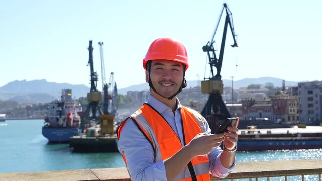 Young asian male engineer wearing a hard hat and safety vest smiling while using a smartphone at a busy commercial shipping port with cranes and cargo ships in the background