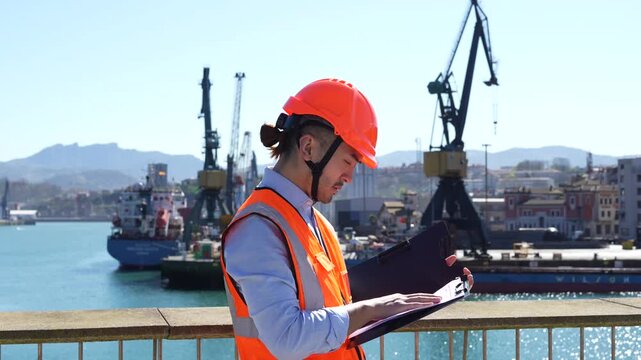 Male dock worker in a safety helmet and vest checking a clipboard while inspecting operations at a busy commercial harbor with cargo ships and cranes in the background on a sunny day