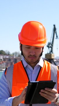 Young asian male engineer in a hard hat and safety vest working on a tablet, managing shipping logistics with a smile at a bustling commercial dockyard under a clear blue sky