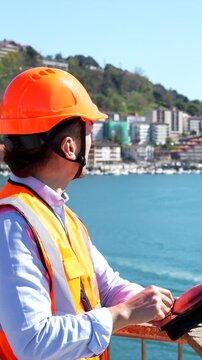 Young asian harbor worker wearing a safety vest and hard hat using a digital tablet while supervising cargo ship operations at the industrial port on a sunny day with the sea in the background