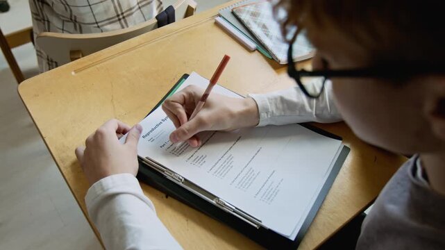 Over-shoulder arc shot of teenage schoolboy writing test on reproductive system at health education lesson, choosing correct answers on question sheet and nodding