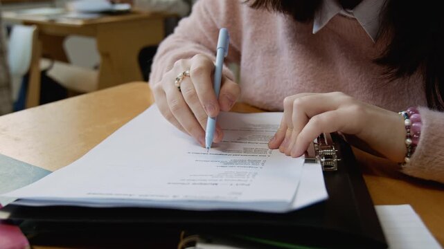 Medium close-up of hands of unrecognizable schoolgirl in pink jumper, with bracelets writing test on pregnancy and reproductive health, contemplating questions and choosing answers on sheet