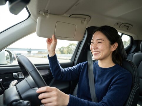 Asian woman driving car adjusts sun visor while smiling in vehicle interior on sunny day