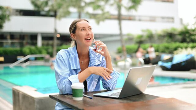 Business woman working remotely on laptop by pool, talking on phone. Digital nomad lifestyle, online work, productivity, travel, luxury resort