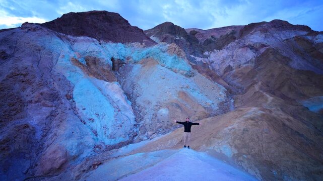 Person stands with outstretched arms on multicolored mineral hills during twilight. Vibrant purple and ochre rock formations create surreal desert landscape. Artist Panel, Death Valley National Park.