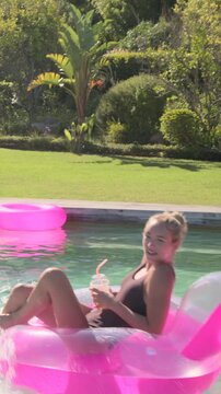Vertical video: Sipping woman in swimsuit on pink float in home pool reacting to spray, copy space