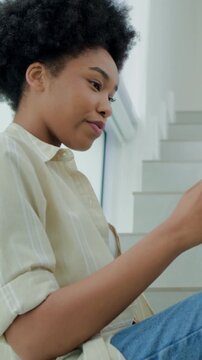 Vertical video: Sipping from glass, woman in cream top and jeans checking phone on stairs by window