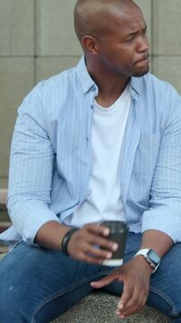 Vertical video: Looking down, speaking, man in blue shirt sipping cup on stone bench reacting