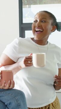 Vertical video: Laughing woman in white tee raising mug at window as friend toasting with pink mug