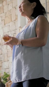 Vertical video: Lifting glass, senior woman in blue top sipping by window, pausing, looking outward
