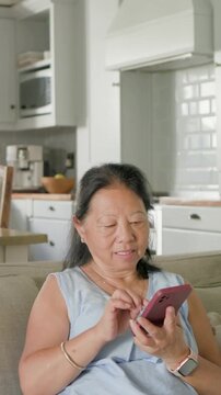 Vertical video: Tapping senior Asian replying to alert on pink phone on couch at home in blue top