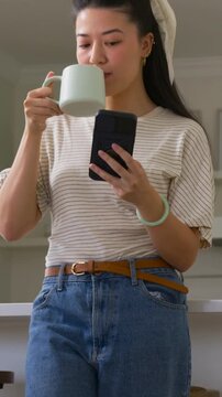 Vertical video: Starting phone, woman sipping mint mug scrolling on black phone at kitchen counter