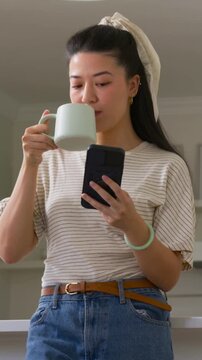 Vertical video: Raising green mug, woman sipping and scrolling phone at home counter to read texts