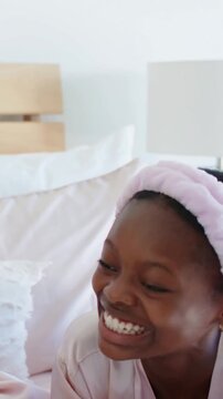 Vertical video: Laughing girl in satin pajamas answering off-camera voice on bed, headband showing