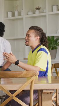 Vertical video: Laughing man in yellow shirt gesturing at kitchen table with friend joining, watch