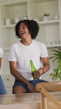 Vertical video: Laughing man in white tee leaning in kitchen, toasting green bottles with friends