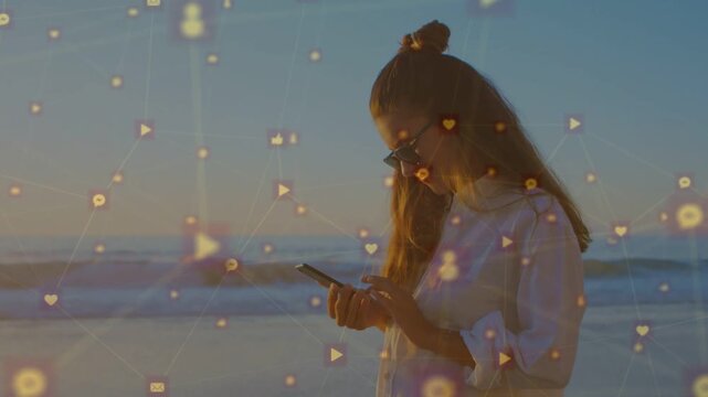 Woman standing on sunset beach tapping phone, checking alerts with social icons growing around her