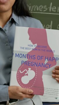 Vertical close-up of midsection and face of young Hispanic female sexual education teacher demonstrating pregnancy guide book at lesson with joyous smile, silhouette of embryo in womb on cover