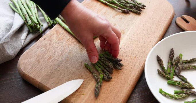 Woman hands cutting fresh green asparagus with sharp white knife on wooden board. Chef preparing raw organic vegetables for healthy vegetarian meal in kitchen. Concept of slicing seasonal ingredients