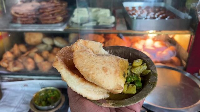A plate of flaky Kachori and spicy potato sabji served with zesty green chilli pickle, with a bustling local shop displaying Jalebi, Samosas, and Gulab Jamun in the background.