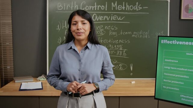 Medium portrait of smiling Hispanic female teacher conducting reproductive health lesson at high school, posing by blackboard with key facts on birth control methods, looking at camera confidently