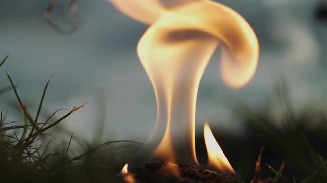 Close-up of a small flame burning in grass against a cloudy sky with warm, golden tones.