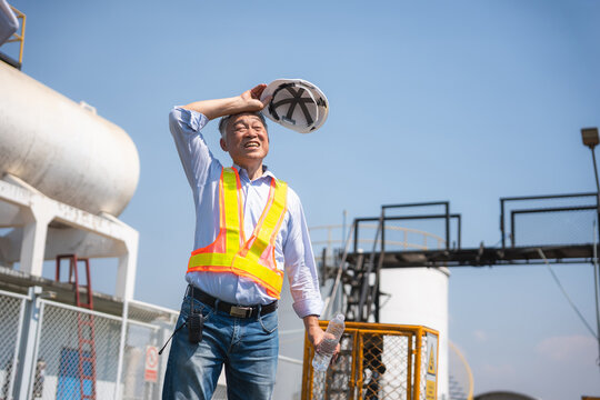 Senior Asian engineer holding helmet and water bottle feeling hot and wiping sweat while working at industrial plant outdoors, Exhausted worker taking a break to drink water during hot sunny day
