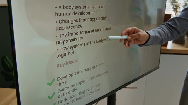 Medium close-up of hand of unrecognizable female teacher demonstrating slide on reproductive system development to class during lesson, highlighting objectives to learn and ideas for discussion