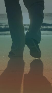 Vertical video: Low sun pushing senior man walking sand toward sea for health, leaving footprints