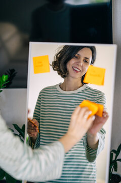 Smiling woman attaching positive affirmation note to mirror indoors