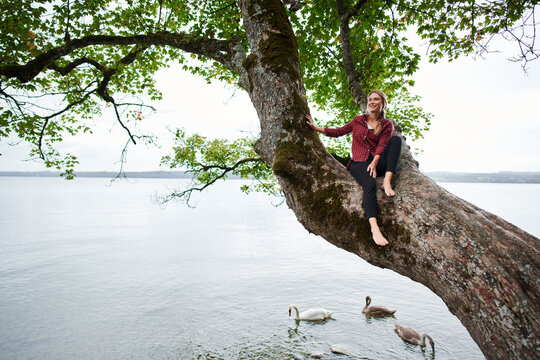 Smiling woman sitting on tree trunk with swans swimming in lake