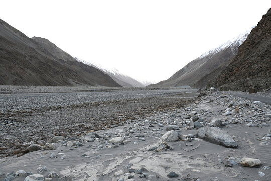 A wide, remote mountain valley landscape in Ladakh, India, with a dry, rocky riverbed and distant snow-capped peaks.