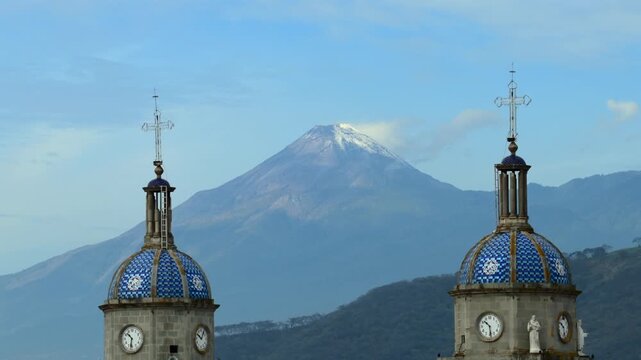 San Juan Bautista church bell towers with blue tiled domes standing in front of the snow capped Colima Volcano in Mexico. Backward aerial