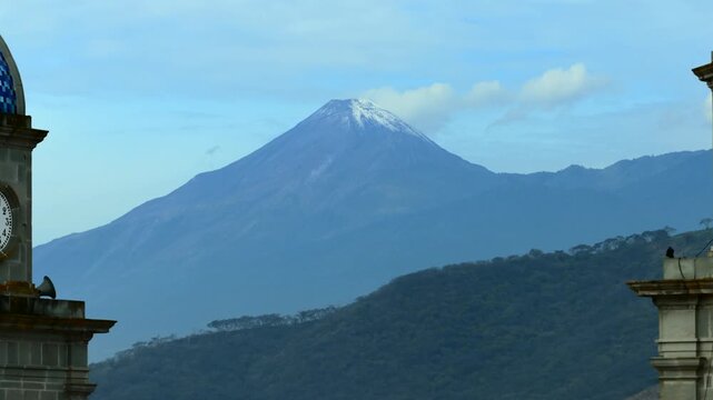 Snow capped Colima Volcano framed between San Juan Bautista church bell towers. Backward aerial reveal shot