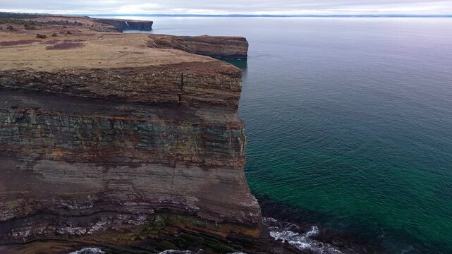 Drone footage revealing layered sedimentary rock cliff face rising above emerald green ocean at Bell Island Newfoundland