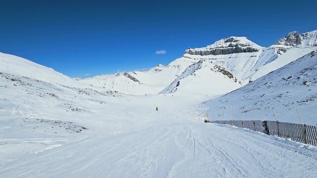 People ski across a snowy Banff mountain run, framed by steep white ridges, crisp blue sky, and expansive Canadian Rockies terrain.