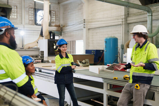 Two workers in yellow workwear discuss CNC machine inspection within bright industrial workshop featuring detailed machinery controls and natural wooden paneling, focused on quality assurance.