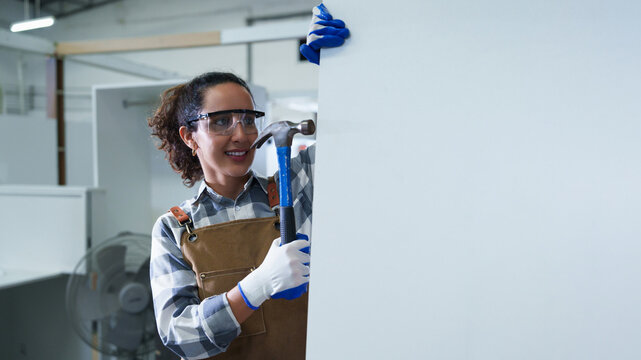 Woman Carpenter Precisely Installing Nail with Hammer on White Wall in Bright Industrial Workshop Featuring Gray Tool Chest, Ventilation Fan, and Precision Woodworking Tools for Industrial Maintenance