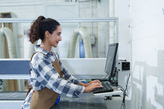 Woman Operating Carpenter CNC Machine with Cutting Equipment in Light-Filled Industrial Factory Setting, Wearing Blue Plaid Work Apron and Monitoring Cutting Process ? Precision Manufacturing in Actio