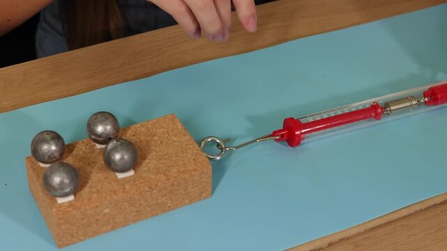 A close up view of a hand attaching a spring scale to a small cork block with lead weights on it.