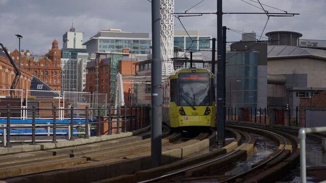 Tram departing on curved tracks in Manchester, UK. Urban skyline with modern and historic buildings, wet rails, and infrastructure highlighting public transport and city mobility.