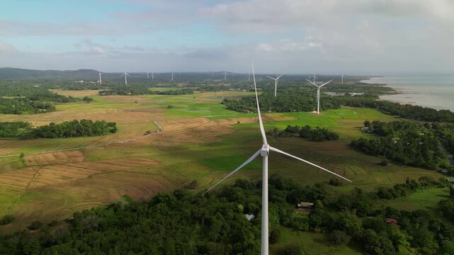 Aerial orbit of a stationary wind turbine overlooking coastal farmland and lush greenery with ocean horizon in Guimaras Island, Philippines