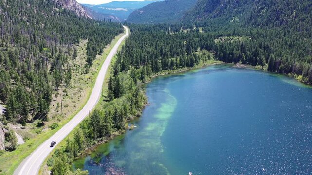 High-angle drone shot of a van traveling on a paved road through rolling hills and green farmland. A cinematic "van life" road trip scene in the BC interior under a bright afternoon sun.