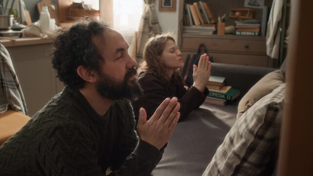 Poor married couple standing on their knees, leaning on couch and praying next to each other, hoping to get through difficult times
