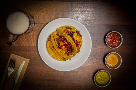 Three Mexican Street Tacos with Cheese, Beans and Salsa Served on a Plate with Beer and Dips