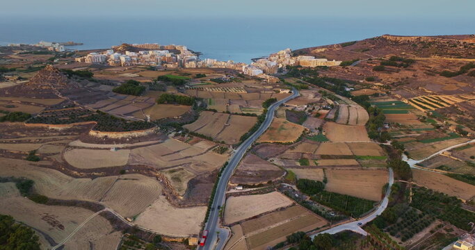 Malta, Comino island: Warm sunlight illuminating the landscape of Gozo, featuring cultivated fields, roads, and hotel buildings coastline in the distance. Aerial view drone flight