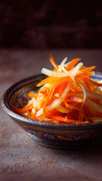 Macro food photography of fresh pickled julienned carrots and daikon radish in an ornate blue and gold ceramic bowl, glistening with vinegar, dramatic side lighting, dark moody background, shallow dep