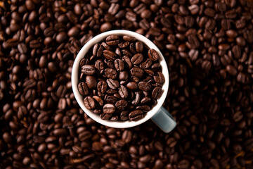 Naklejka premium Top down view of a white ceramic cup filled with aromatic dark roasted coffee beans on a background of scattered beans for cafe and breakfast concept